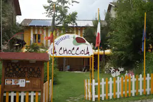 Schild „La Choccola“ am Campingplatz mit Restaurant-Eingang, Fahnen, weißem Zaun und Rasen im Garten.