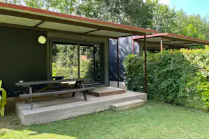 Grüne Campingunterkunft mit Holzterrasse, Sitzbank und Tisch, überdachtem Eingang und Hecken im Garten.