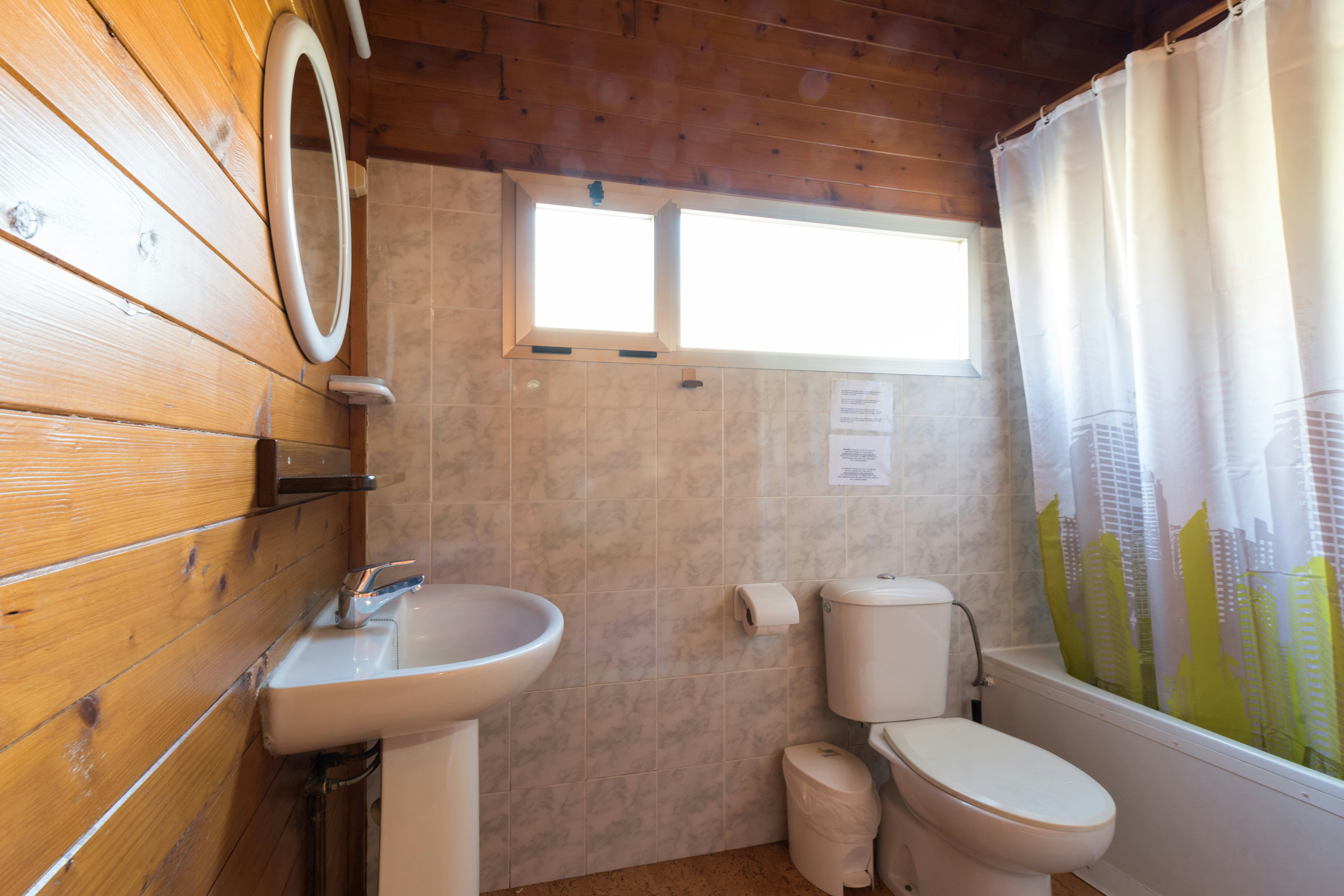 Modernes Badezimmer in einem Holzhaus mit Waschbecken, Toilette, Dusche und Vorhang, hellen Fliesen und Wandpaneelen.