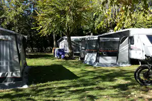 Wohnwagen und Zelte auf einem grünen Campingplatz, daneben ein Fahrrad und Hecken mit Bäumen im Hintergrund.