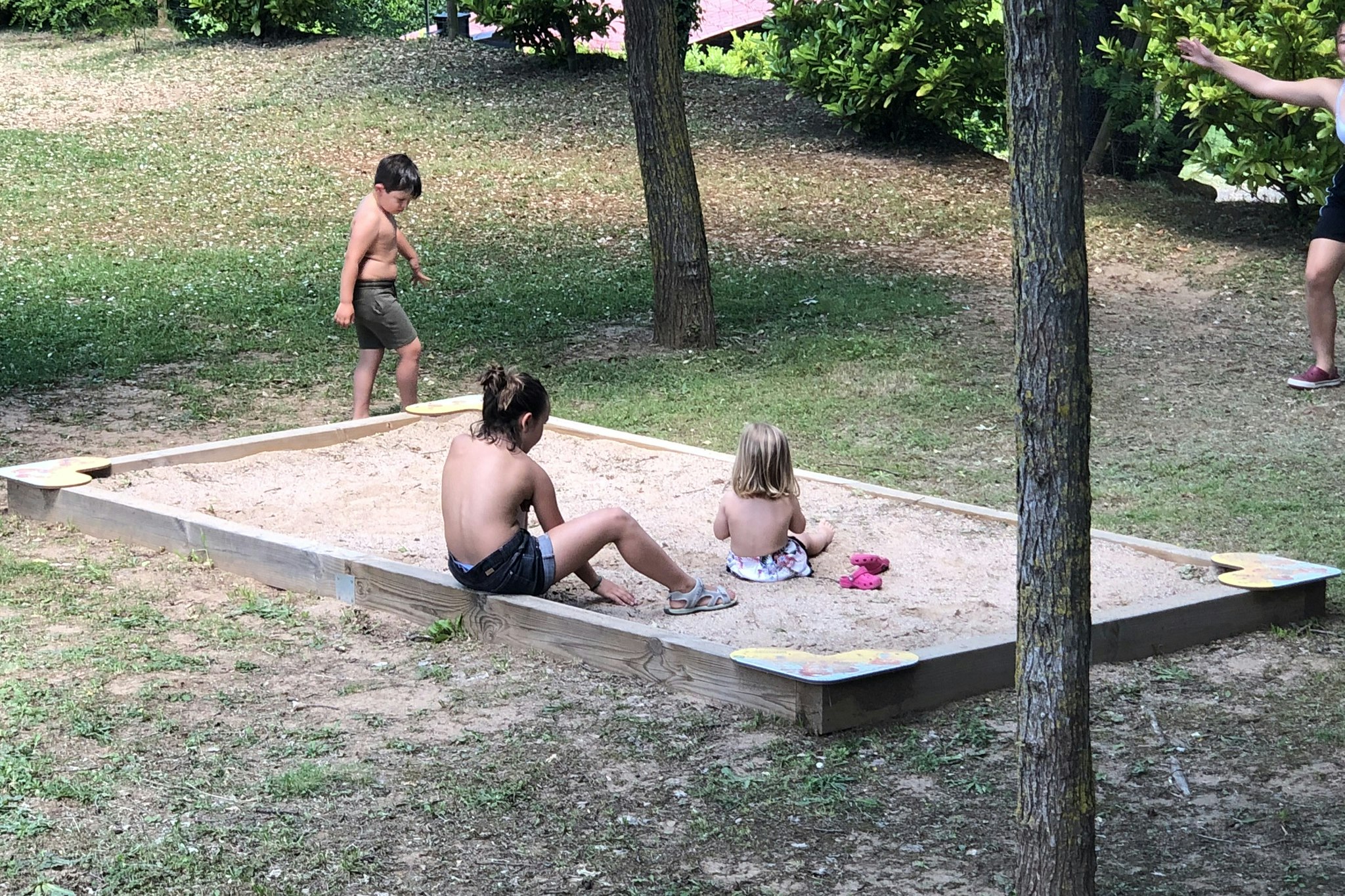 Kinder spielen in einer Sandkiste auf dem Campingplatz, umgeben von Rasen und Bäumen.