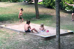 Kinder spielen in einer Sandkiste auf dem Campingplatz, umgeben von Rasen und Bäumen.