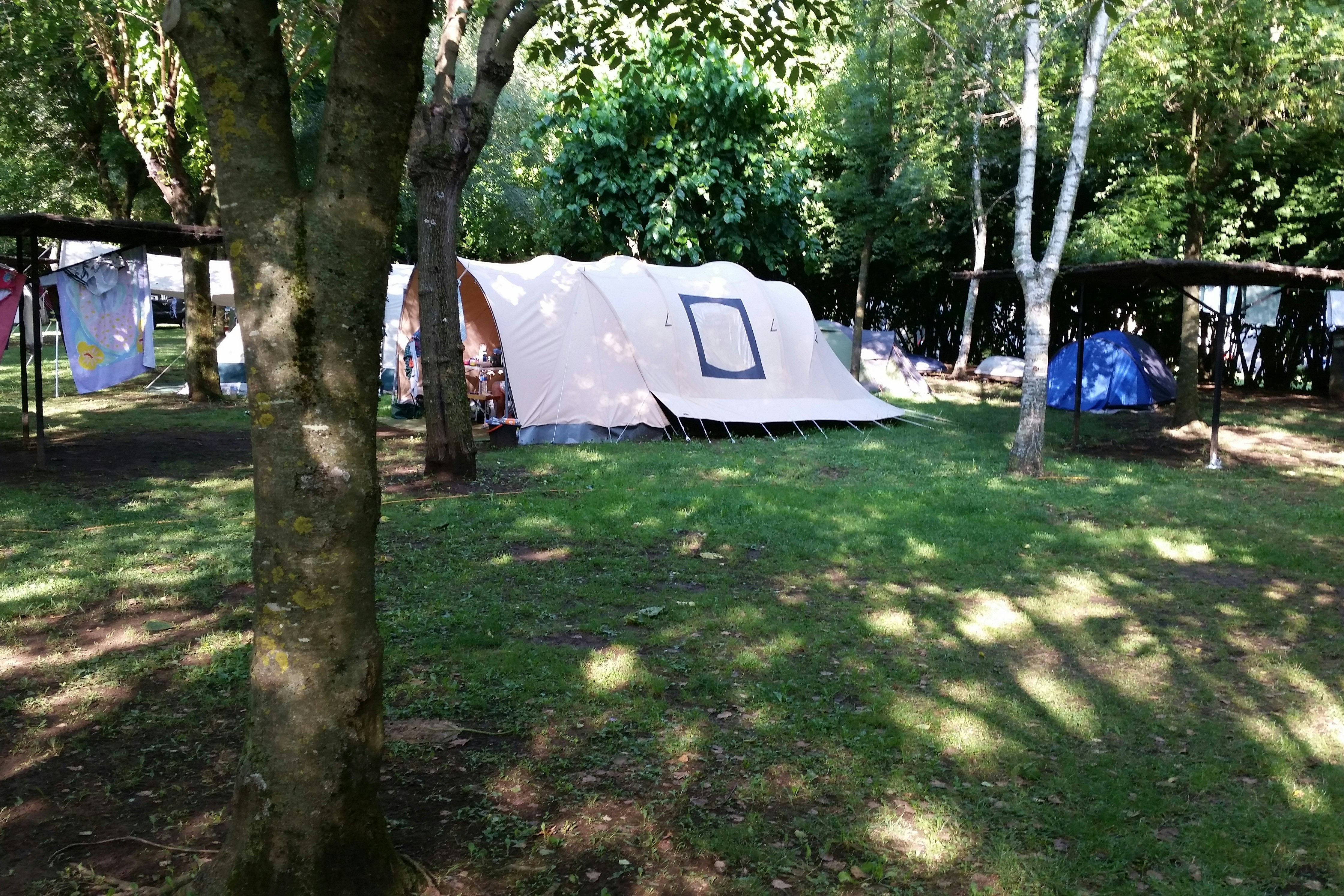 Weißes Zelt auf einem Campingplatz im Grünen zwischen Bäumen, mit weiteren Zelten im Hintergrund.