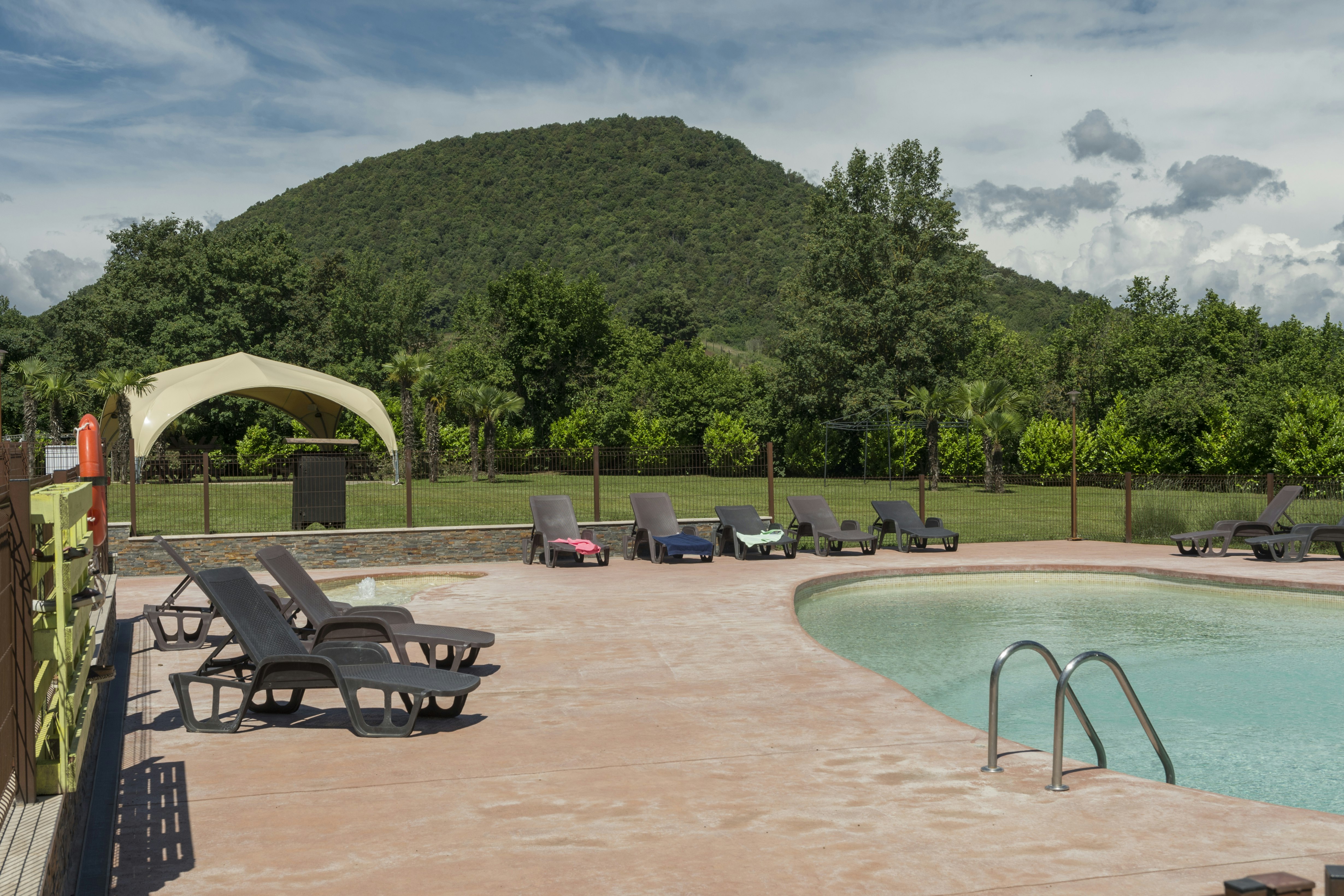 Camping-Pool mit eingelassenem Becken, mehreren Liegen und Sonnenschirm, im Hintergrund Bäume und ein bewaldeter Hügel.