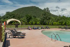 Camping-Pool mit eingelassenem Becken, mehreren Liegen und Sonnenschirm, im Hintergrund Bäume und ein bewaldeter Hügel.