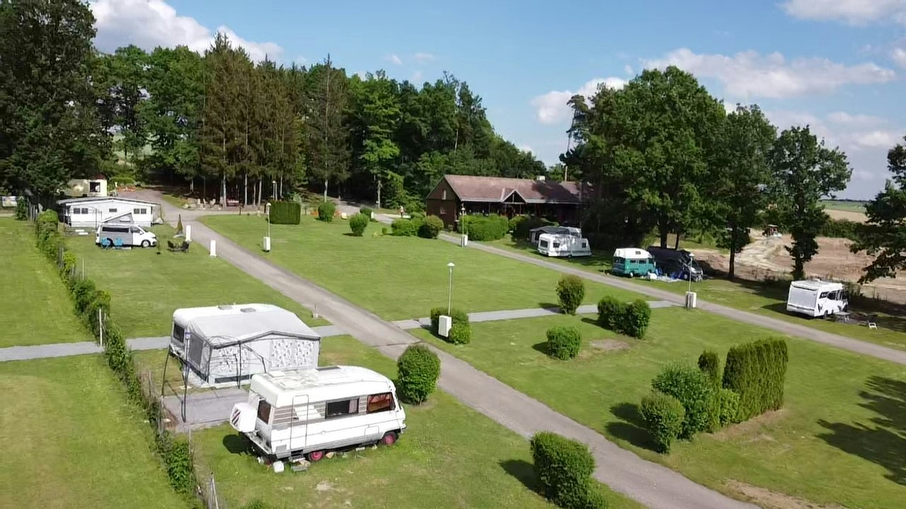 Campingplatz mit Wohnwagen auf Stellplätzen im Grünen, gepflasterte Wege, Hecken und Bäume, im Hintergrund Gebäude und Wiese.