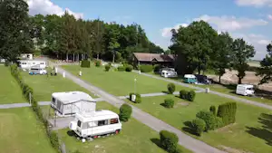 Campingplatz mit Wohnwagen auf Stellplätzen im Grünen, gepflasterte Wege, Hecken und Bäume, im Hintergrund Gebäude und Wiese.