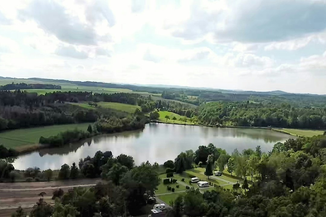 Blick auf einen See mit Uferwald und grünen Hügeln, darunter ein Campingplatz mit Stellplätzen und Wegen.