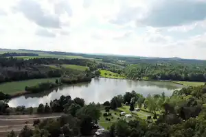 Blick auf einen See mit Uferwald und grünen Hügeln, darunter ein Campingplatz mit Stellplätzen und Wegen.