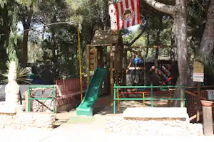 Spielplatz mit grüner Rutsche auf Holzpodest, umgeben von Bäumen und Zäunen, im Campingbereich.