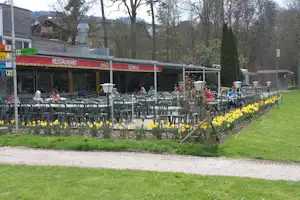 Restaurant am Campingplatz mit Terrasse, vielen Stühlen und Tischen, Blumenbeeten und Rasen im Garten.