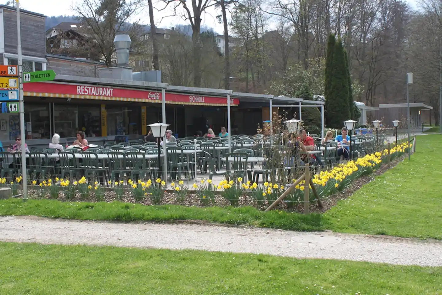 Restaurant am Campingplatz mit Terrasse, vielen Stühlen und Tischen, Blumenbeeten und Rasen im Garten.