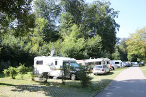 Wohnmobile und Autos auf einem Campingplatz, gepflasterter Weg und viel Grün mit hohen Bäumen im Hintergrund.