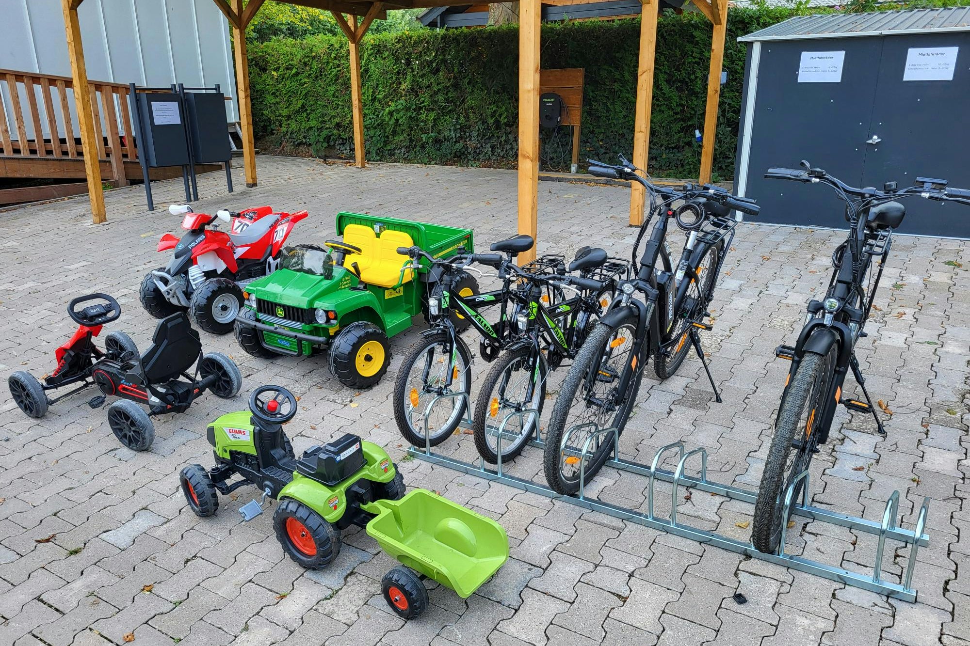 Fahrräder und Kinderfahrzeuge auf einem gepflasterten Platz vor einem Schuppen, mit Hecken im Hintergrund.