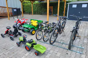 Fahrräder und Kinderfahrzeuge auf einem gepflasterten Platz vor einem Schuppen, mit Hecken im Hintergrund.