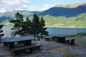 Steintisch und Bänke am Campingplatz mit Blick auf einen See und grüne Berge im Hintergrund.