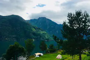 Zelte auf einer grünen Wiese am See, im Hintergrund bewaldete Berge und Bäume unter wolkigem Himmel.