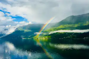Regenbogen über einem See mit Spiegelung, grünen Berghängen und Wolken am Himmel.