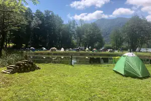 Grünes Zelt auf einer Campingwiese am Teich, im Hintergrund weitere Zelte und Bäume vor den Bergen.