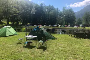 Grünes Campingzelt auf einer Wiese, daneben Tisch und Stühle, im Hintergrund weitere Zelte und Fahrräder am Zaun.