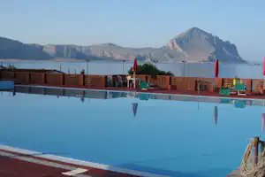 Campingpool mit blauem Wasser, Ziegelmauer und Liegen am Beckenrand, im Hintergrund Berge und See.