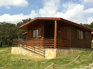 Holzbungalow mit Terrasse, Geländer und großen Fenstern auf grünem Rasen, im Hintergrund Bäume und ein Weg.