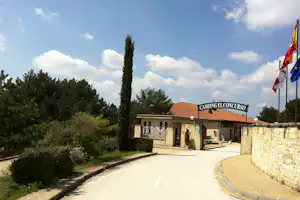 Eingang vom Campingel Concorso mit Steinmauer, Zufahrtsweg, Fahnen und Gebäude mit roten Ziegeldach im Grünen.