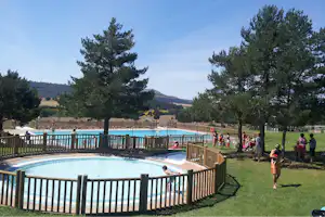 Schwimmbecken am Campingplatz mit Holzgeländer, Bäumen und Blick auf grüne Hügel im Hintergrund.