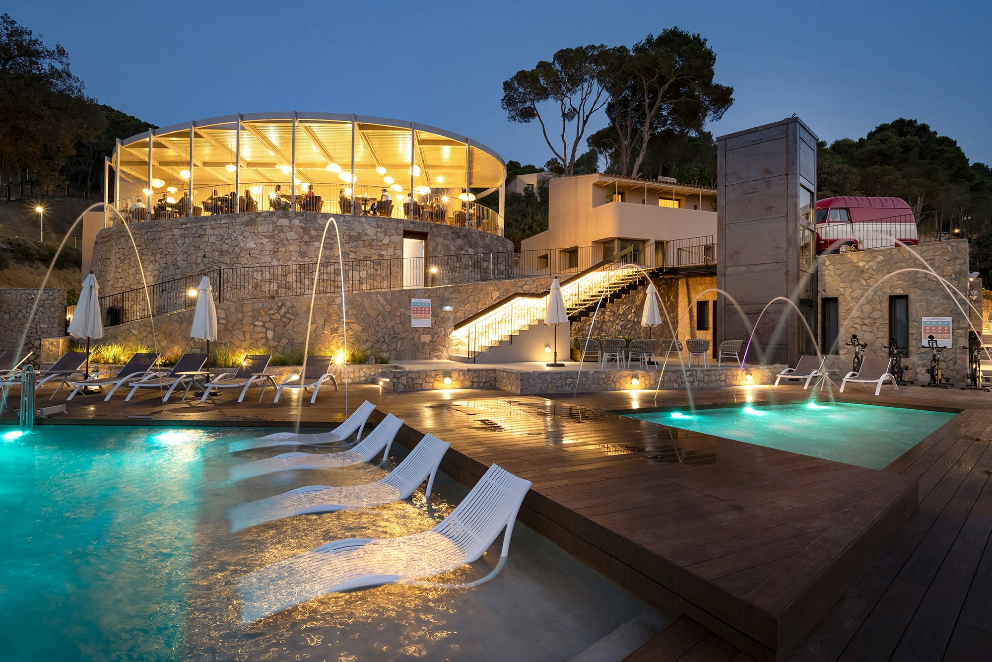 Beleuchteter Camping-Pool mit Liegen am Rand, moderne Gebäude und Steinmauer im Hintergrund bei Nacht.