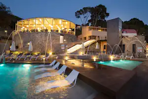 Beleuchteter Camping-Pool mit Liegen am Rand, moderne Gebäude und Steinmauer im Hintergrund bei Nacht.