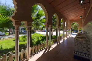 Veranda mit Säulen und Fliesenboden, Blick auf Palmen und gepflegten Garten am Campingplatz, im Hintergrund Fahrzeuge.