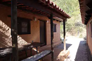 Holzhütte mit Veranda und Bank, Steinwand und Fenster, Blick auf den Weg zwischen Gebäuden im Grünen.