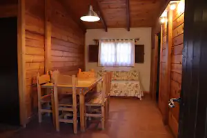Holzblockhütte innen: Esstisch mit Stühlen, gepolsterte Bank und Fenster mit Vorhang, warme Beleuchtung an der Decke.