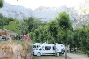 Wohnmobile und Campingausrüstung am Stellplatz, umgeben von Bäumen, mit Blick auf hohe Felsberge im Hintergrund.