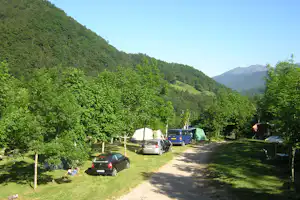 Campingplatz mit Autos und Zelten auf einer Wiese, umgeben von Bäumen und Bergen im grünen Tal.