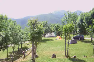 Campingwiese mit drei Zelten, Bäumen und geparktem Auto am Rand, im Hintergrund Berge und bewaldete Hänge.
