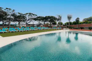 Schwimmbecken mit türkisfarbenem Wasser, Reihe blauer Liegen am Rand, gepflegter Rasen und Bäume im Hintergrund.