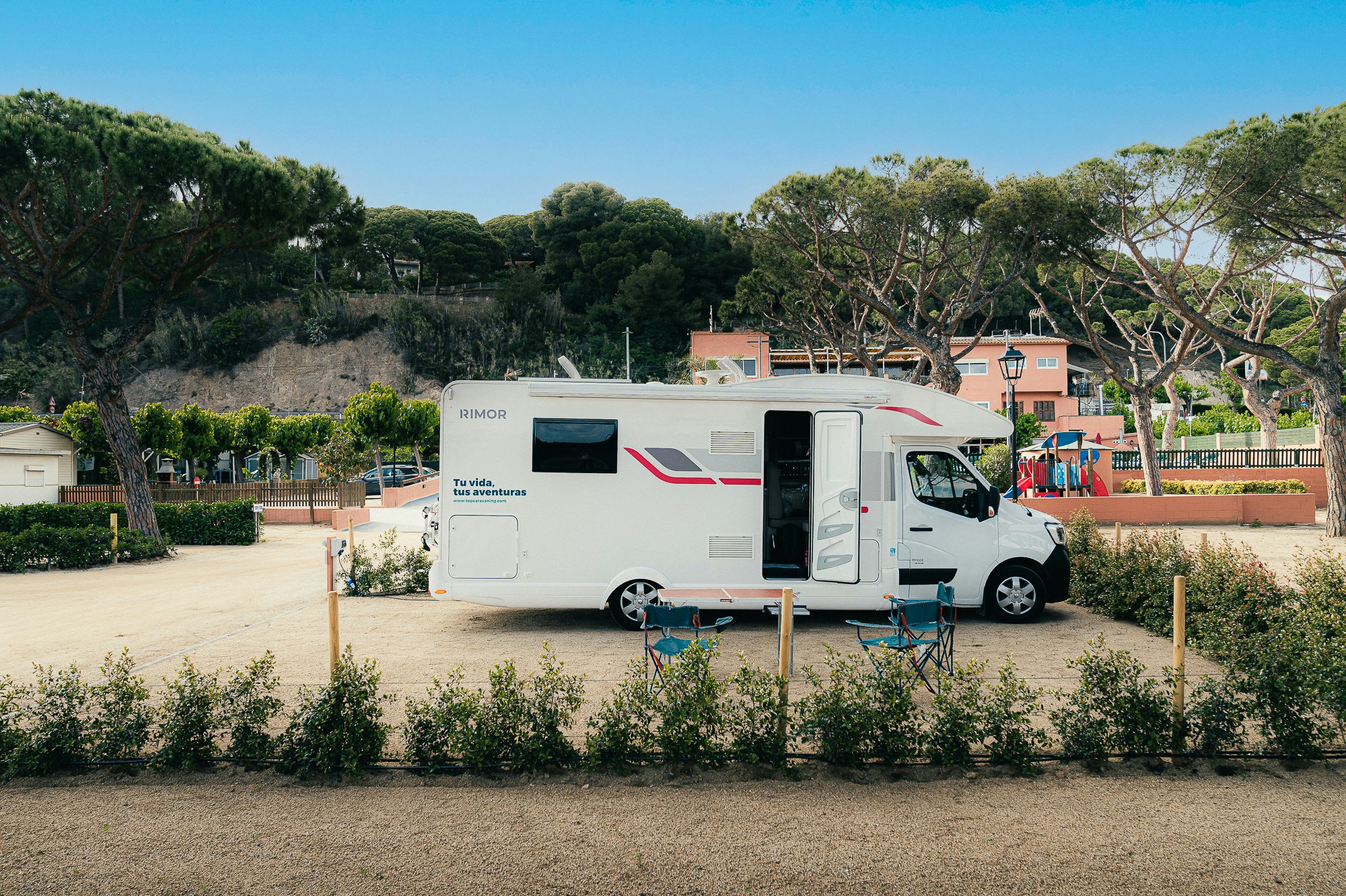 Weißes Wohnmobil auf einem Stellplatz im Campinggelände, umgeben von Bäumen und einem Weg, im Hintergrund Gebäude.