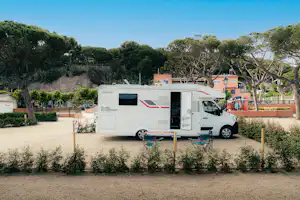 Weißes Wohnmobil auf einem Stellplatz im Campinggelände, umgeben von Bäumen und einem Weg, im Hintergrund Gebäude.