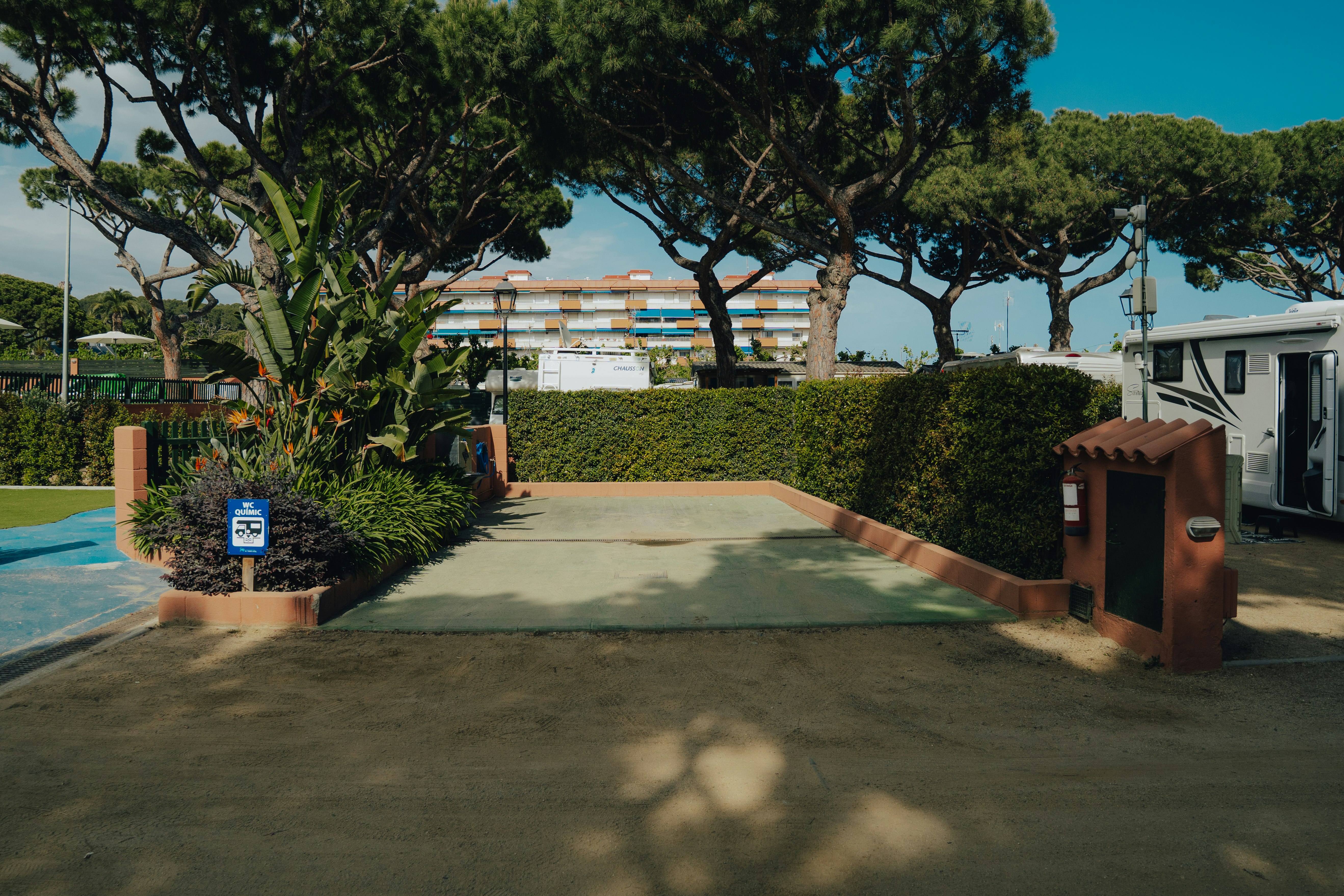 Eingang zum Campingplatz mit gepflastertem Weg, Hecken und Bäumen, daneben ein Gebäude und ein kleiner Brunnen.