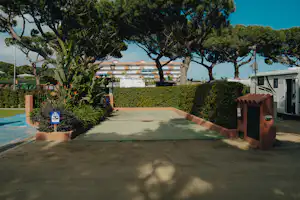Eingang zum Campingplatz mit gepflastertem Weg, Hecken und Bäumen, daneben ein Gebäude und ein kleiner Brunnen.