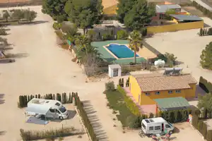 Luftaufnahme eines Campingplatzes mit Pool, Wohnmobilen auf Stellplätzen und Wegen zwischen Bäumen und kleinen Gebäuden.