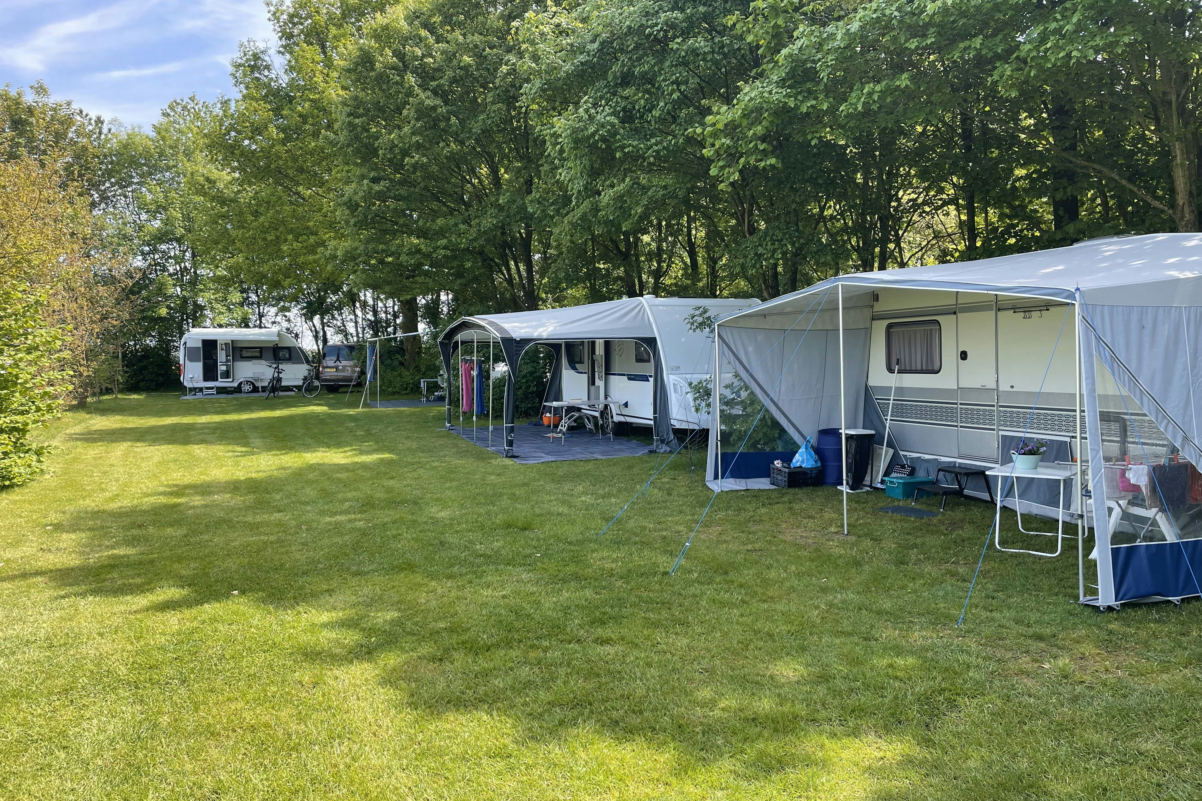 Wohnwagen mit Vorzelt und Campingmöbeln auf dem Gras, daneben weitere Wohnmobile und Fahrräder vor Bäumen.
