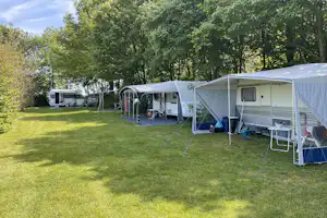 Wohnwagen mit Vorzelt und Campingmöbeln auf dem Gras, daneben weitere Wohnmobile und Fahrräder vor Bäumen.