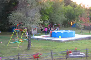 Kinder spielen am blauen Planschbecken auf dem Campingplatz, im Hintergrund Zelte, Bäume und eine Picknick-Tafel.
