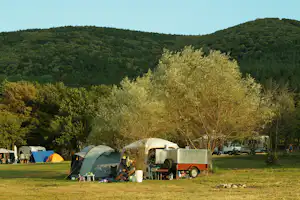 Campingplatz mit mehreren Zelten, Wohnwagen und einem Anhänger auf einer Wiese, umgeben von Bäumen und Hügeln.