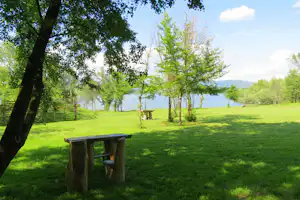 Grüne Campingwiese am See mit Holztisch und Bänken unter Bäumen, im Hintergrund Wasser und Hügel.