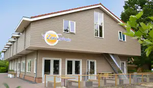 Ferienhaus mit Balkon und großen Fenstern, Holzverkleidung, Sitzbereich und Weg am Campingplatz, im Hintergrund Bäume.