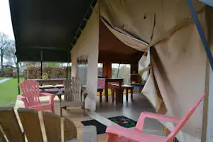 Campingzelt mit Liegestühlen und Holztisch im Innenbereich, seitlich offene Plane, Rasen und Hecken im Hintergrund.