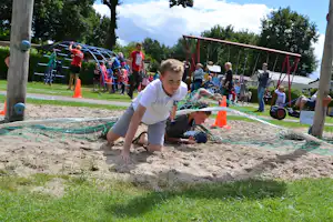 Kinder spielen im Sand auf dem Campingplatz, im Vordergrund ein Junge mit Netz und orangefarbenen Markierungen.